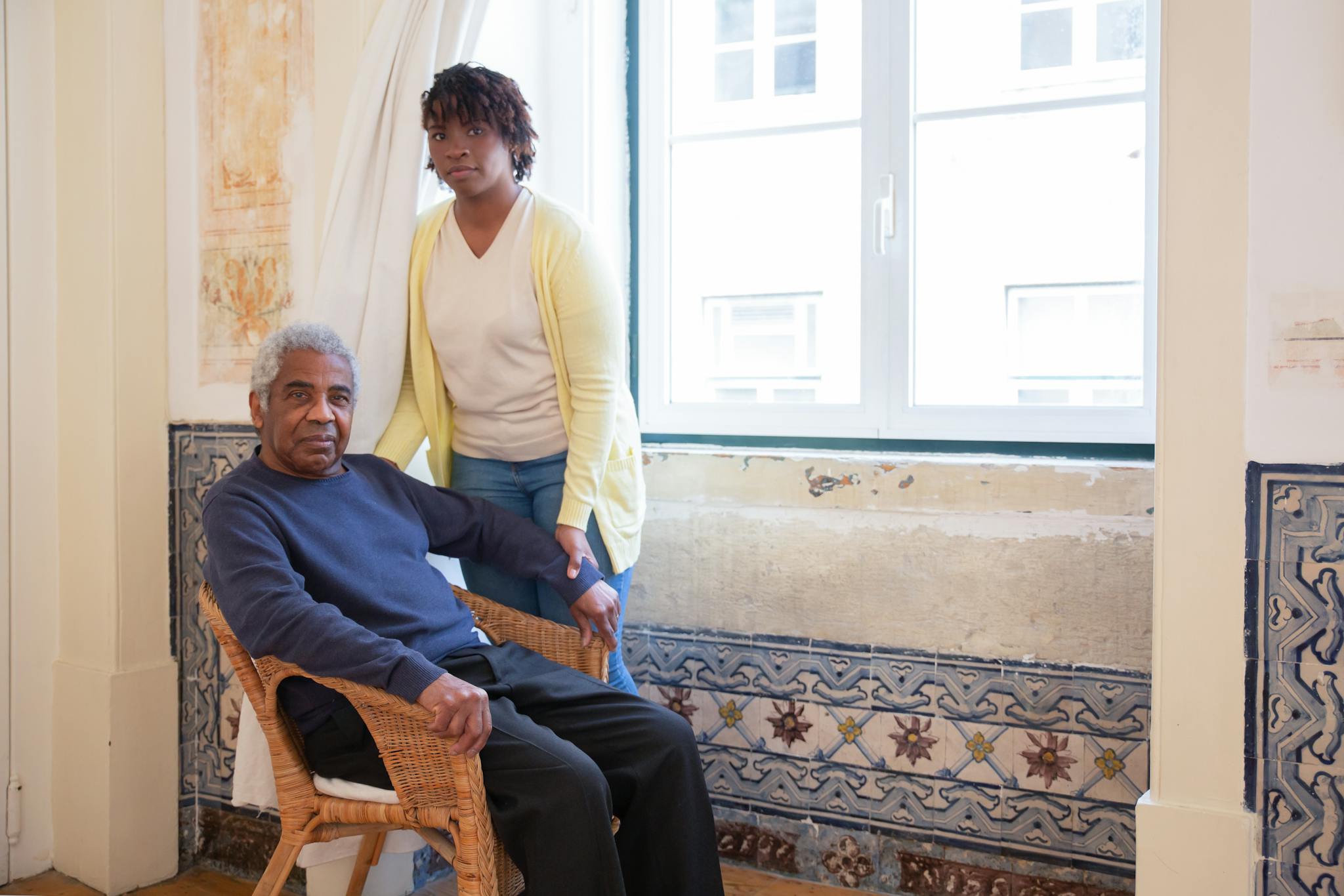 About Us Elderly man seated with caregiver standing beside him at home in Portugal.