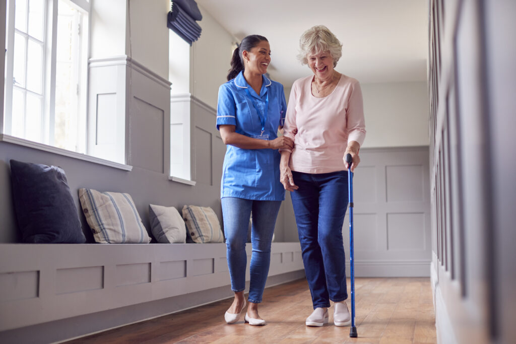 Home senior woman at home using walking stick being helped by female care worker in uniform