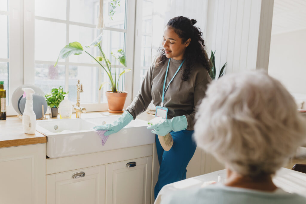 Holistic Care rare view of gray haired lady talking to black helping volunteer standing next to kitchen sink in blue raisin gloves, cleaning room carefully using spray and microfiber cloth, wearing badge with name